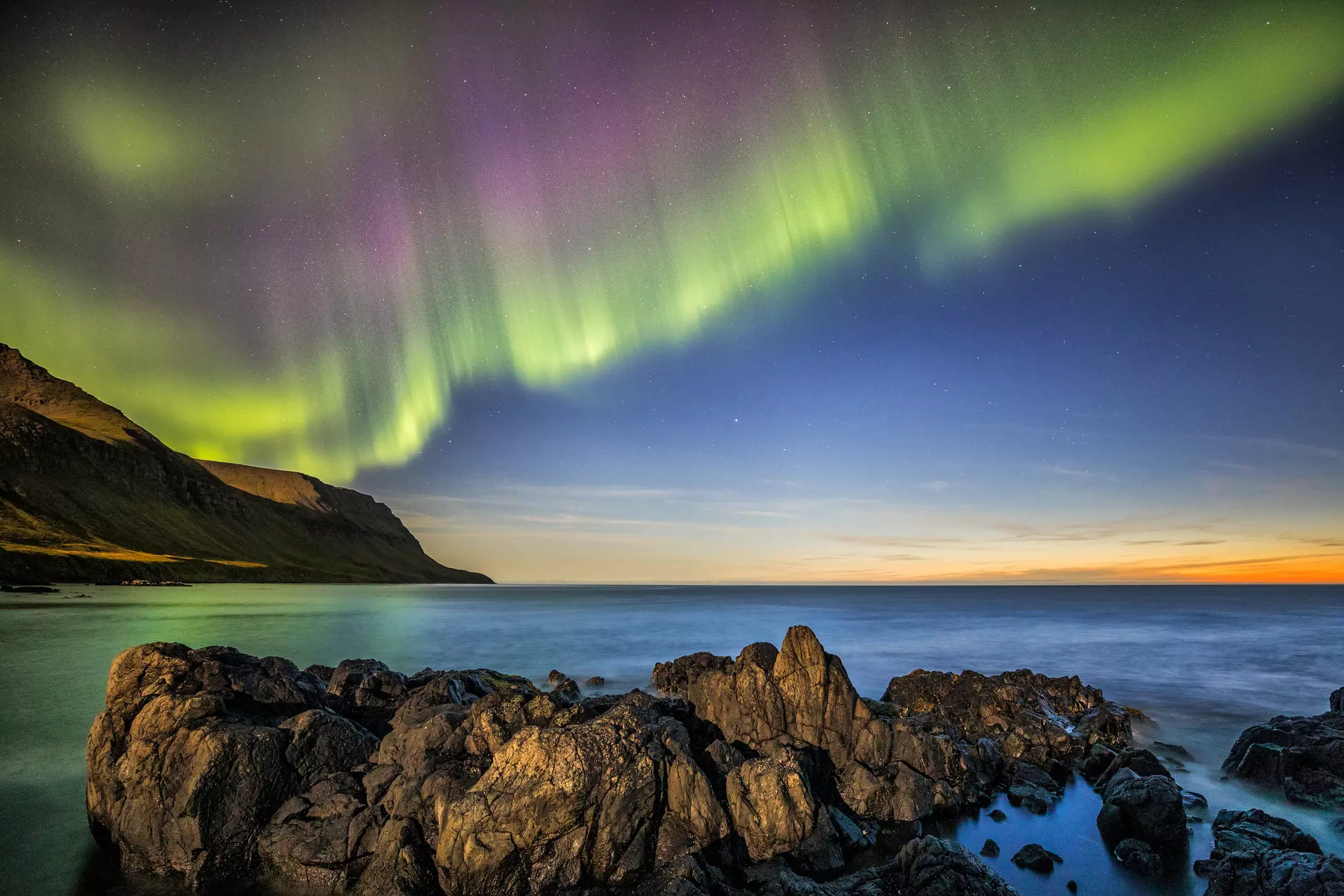 The northern lights over a hill and sea are visible with sunlight still on the edge of the horizon; there are rocks in the foreground.