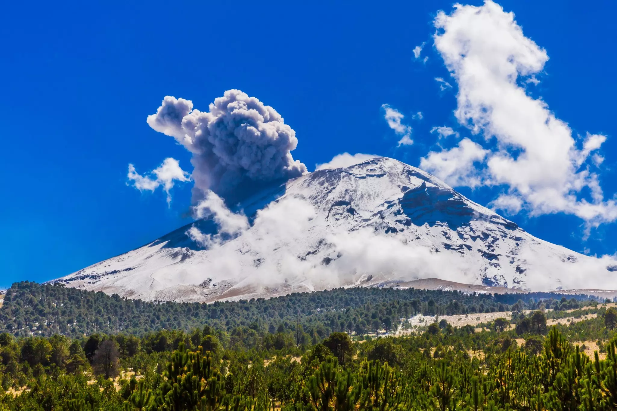 A wide view of a volcano covered with snow and releasing smoke on a sunny day.