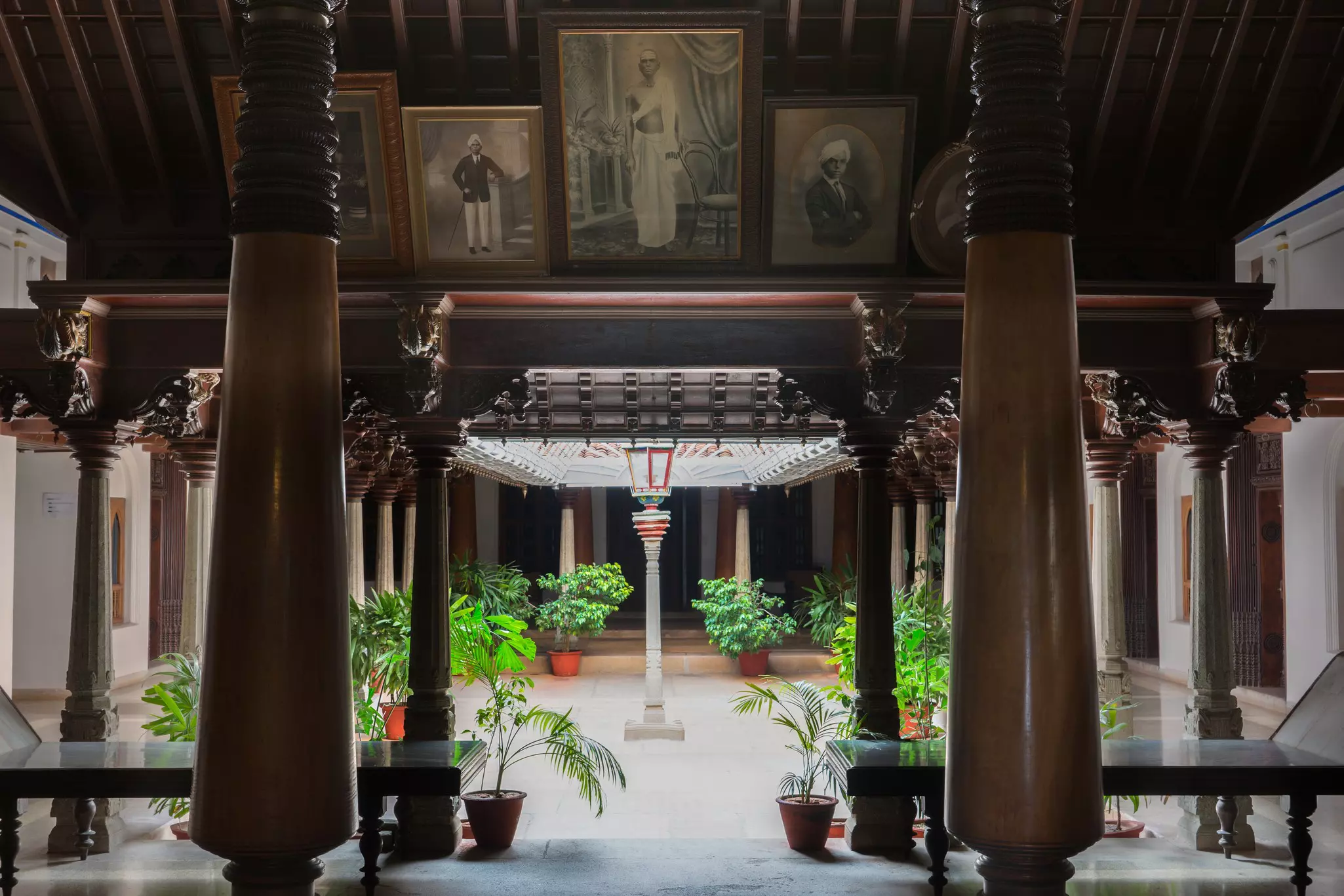  The entrance hall of a mansion leads to an open -air courtyard. Old sepia photos, green plants, dark brown wood are pictured.