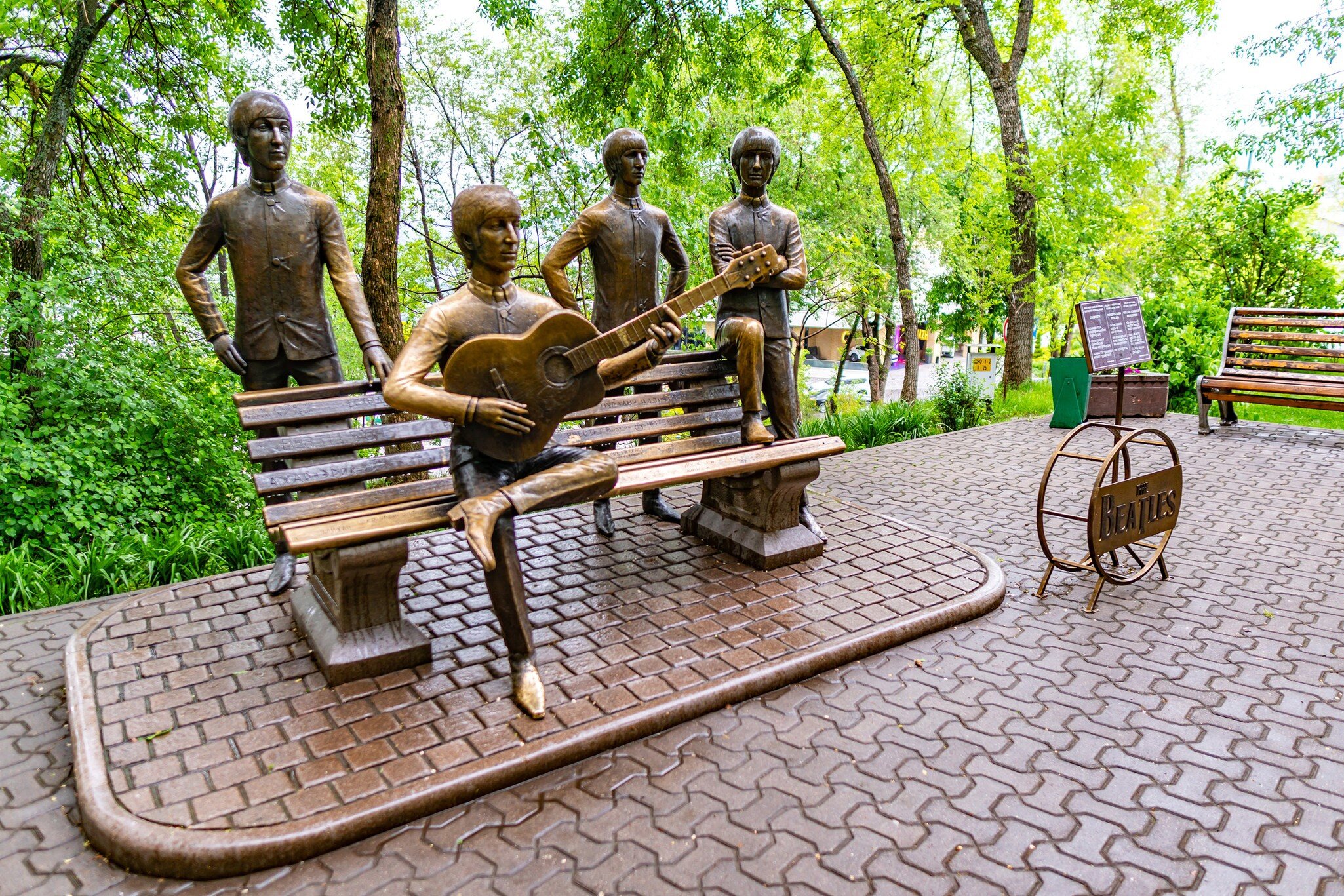Four bronze figures around a park bench, one playing guitar, with trees and a paved plaza behind