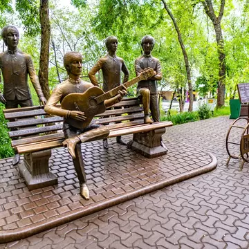 Four bronze figures around a park bench, one playing guitar, with trees and a paved plaza behind