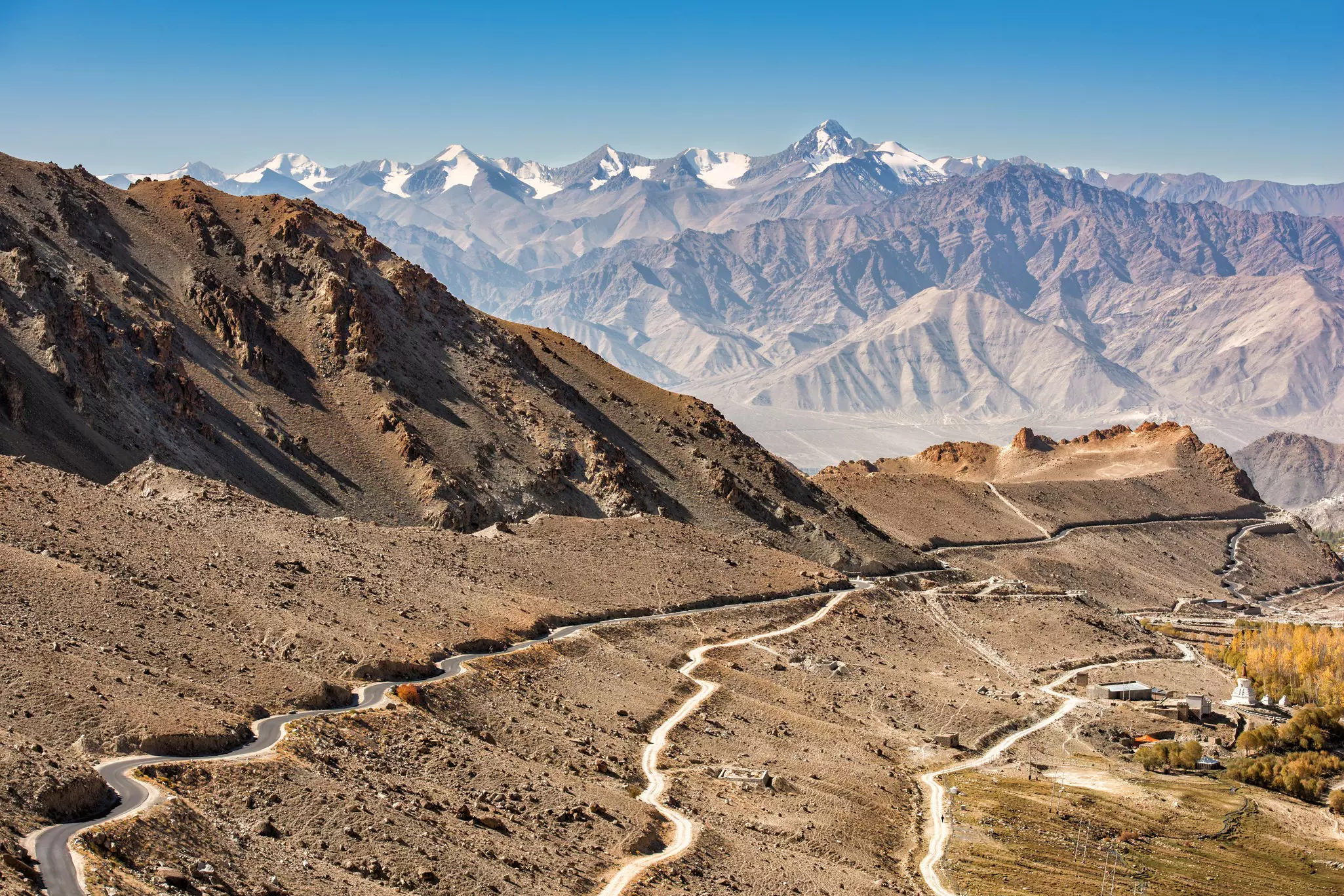 A mountain road weaves around bends and up and down hills overlooked by snow-capped peaks.
