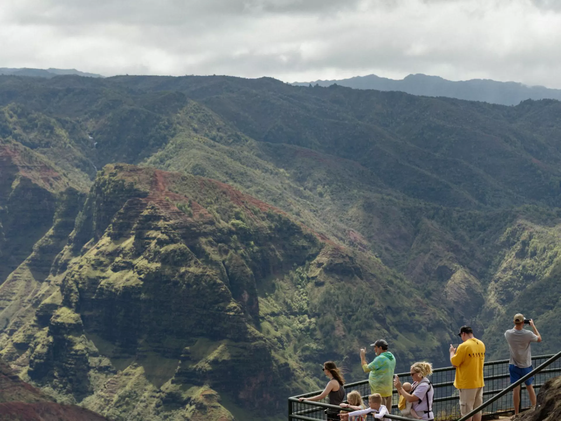 People look and take pictures of a large green canyon in Hawaiʻi.