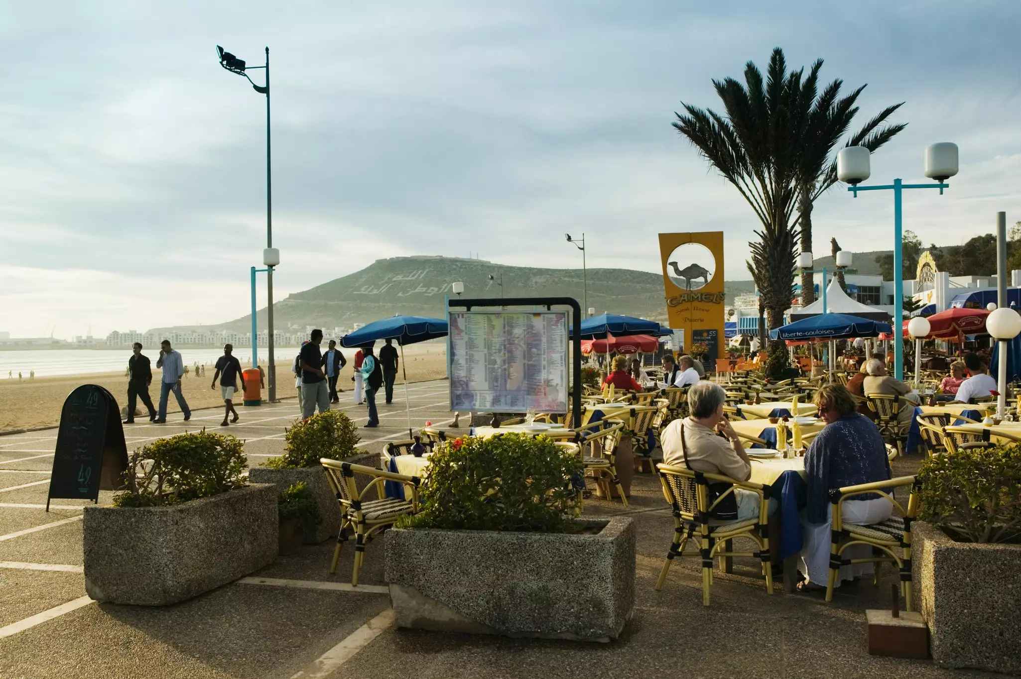 People sit at a cafe and walk along a promenade by a beach in Morocco.