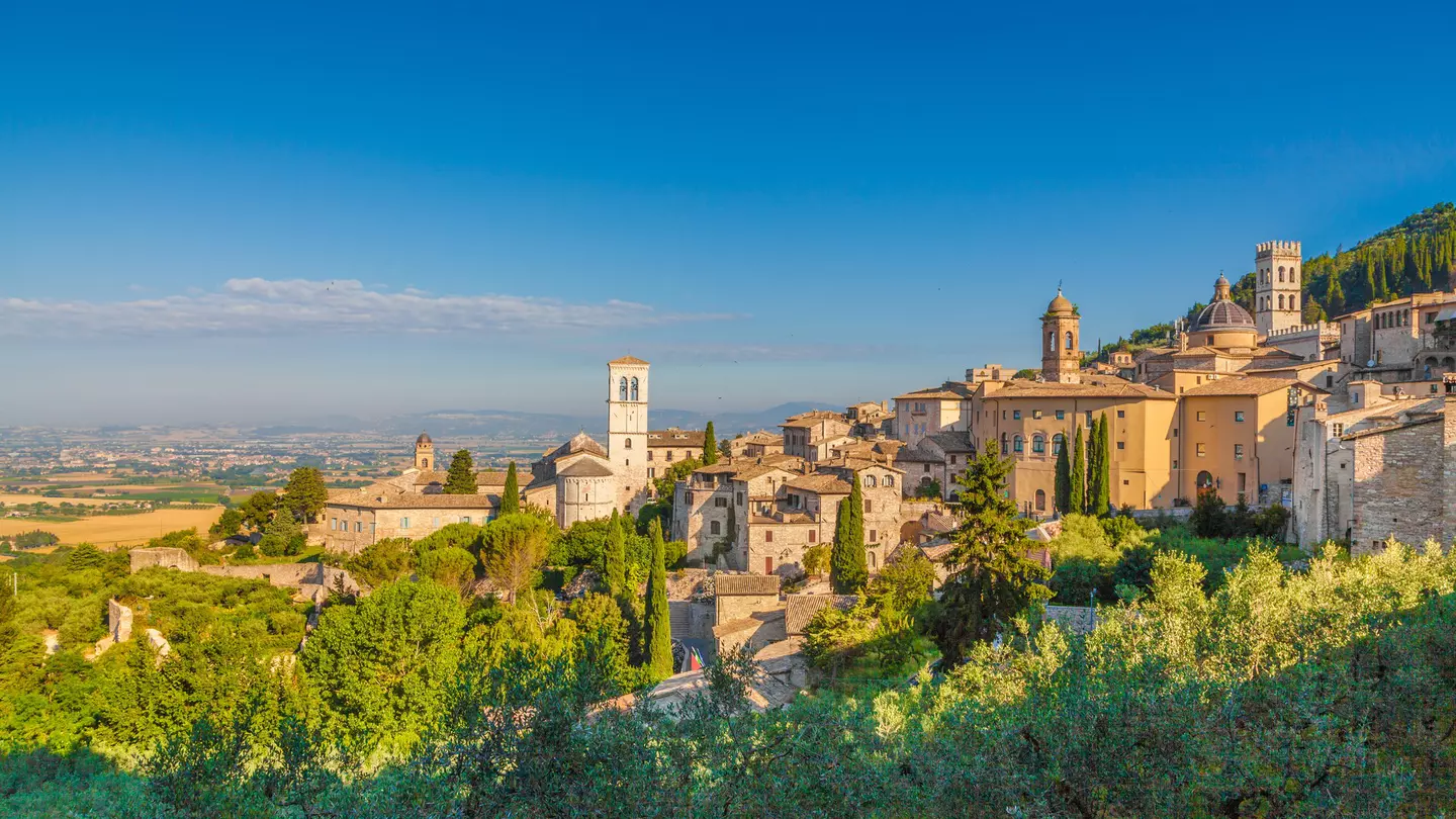 The sandstone-coloured buildings of Assisi glow in the sunshine, with a clear blue sky behind and countryside in the distance