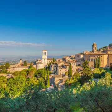 The sandstone-coloured buildings of Assisi glow in the sunshine, with a clear blue sky behind and countryside in the distance