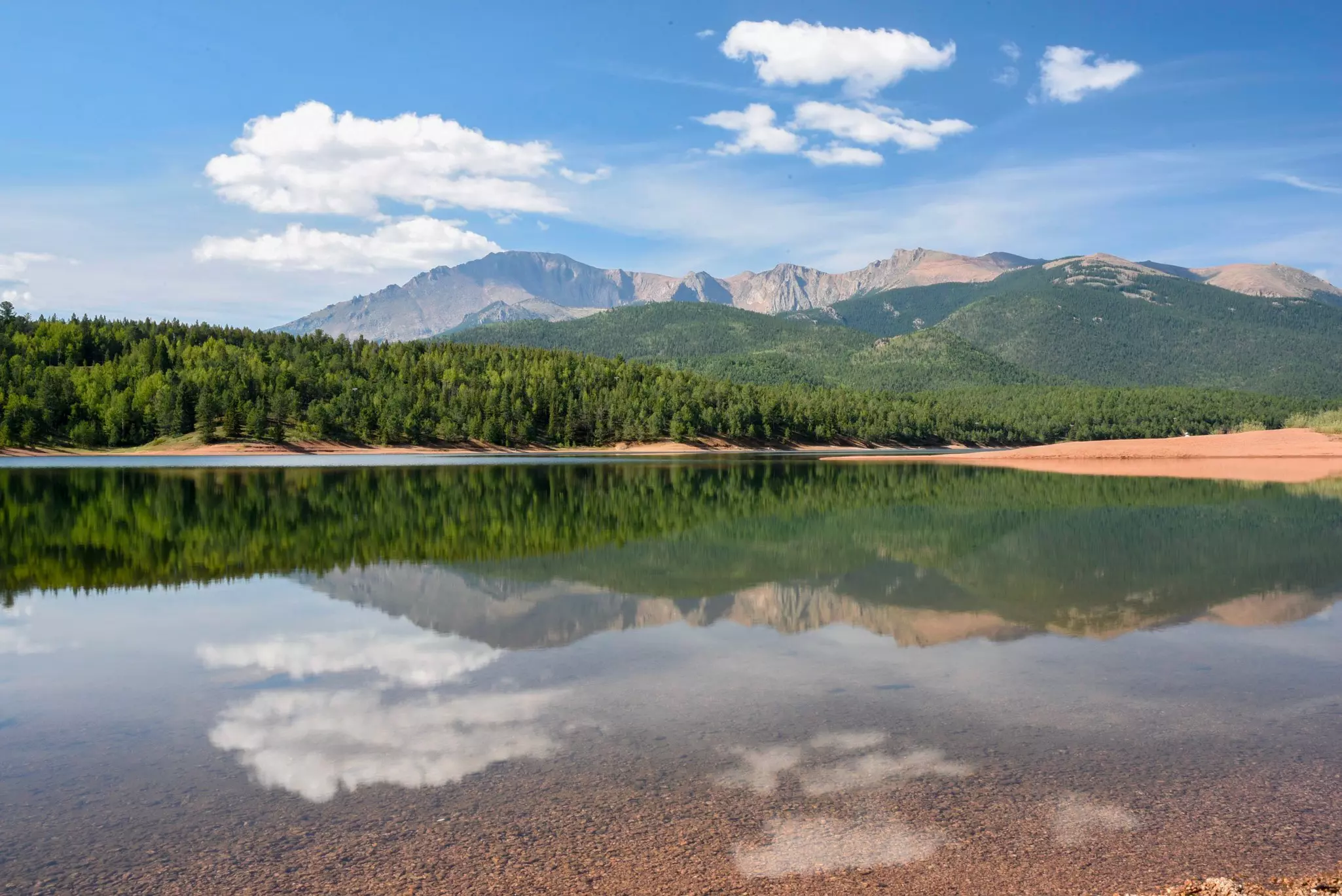 A blue sky and clouds are reflected in a still mountain lake. Pine trees are visible along the shore, and mountains can be seen in the distance.