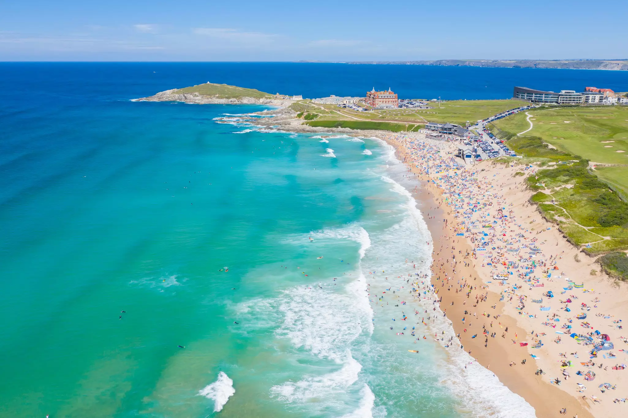 Waves crash on a vast sandy beach that is packed with people enjoying a sunny day. A large building dominates a peninsula of land in the distance.