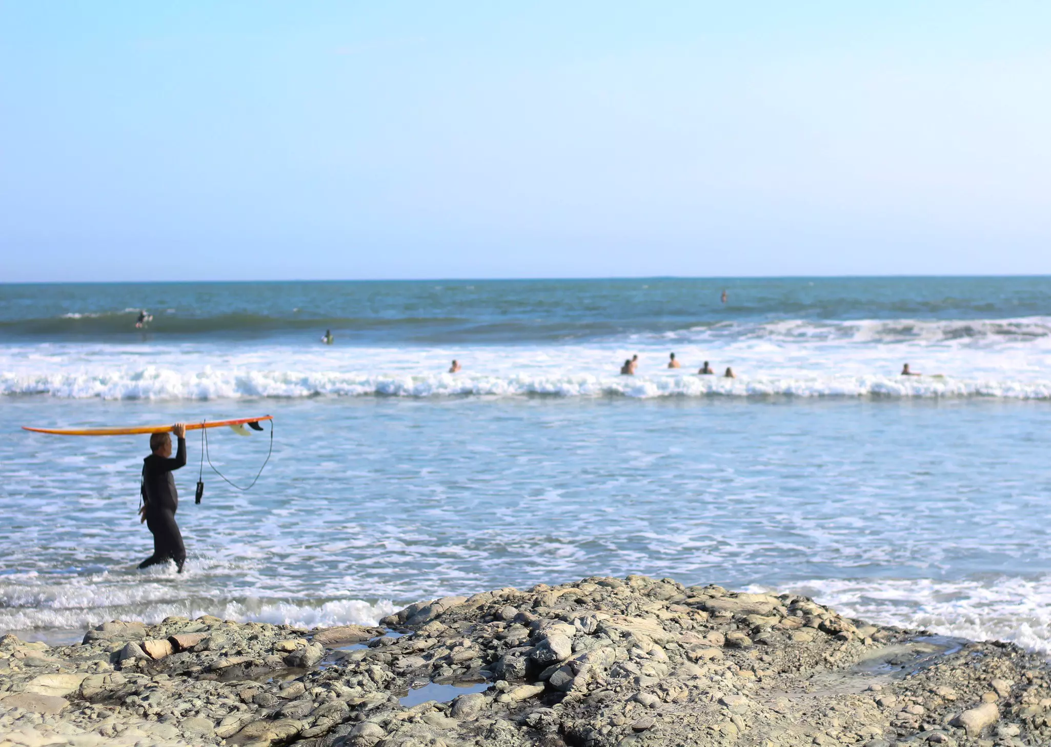 Man in black wetsuit carrying a surfboard above his head walking into the surf with swimmers and the ocean in the background.