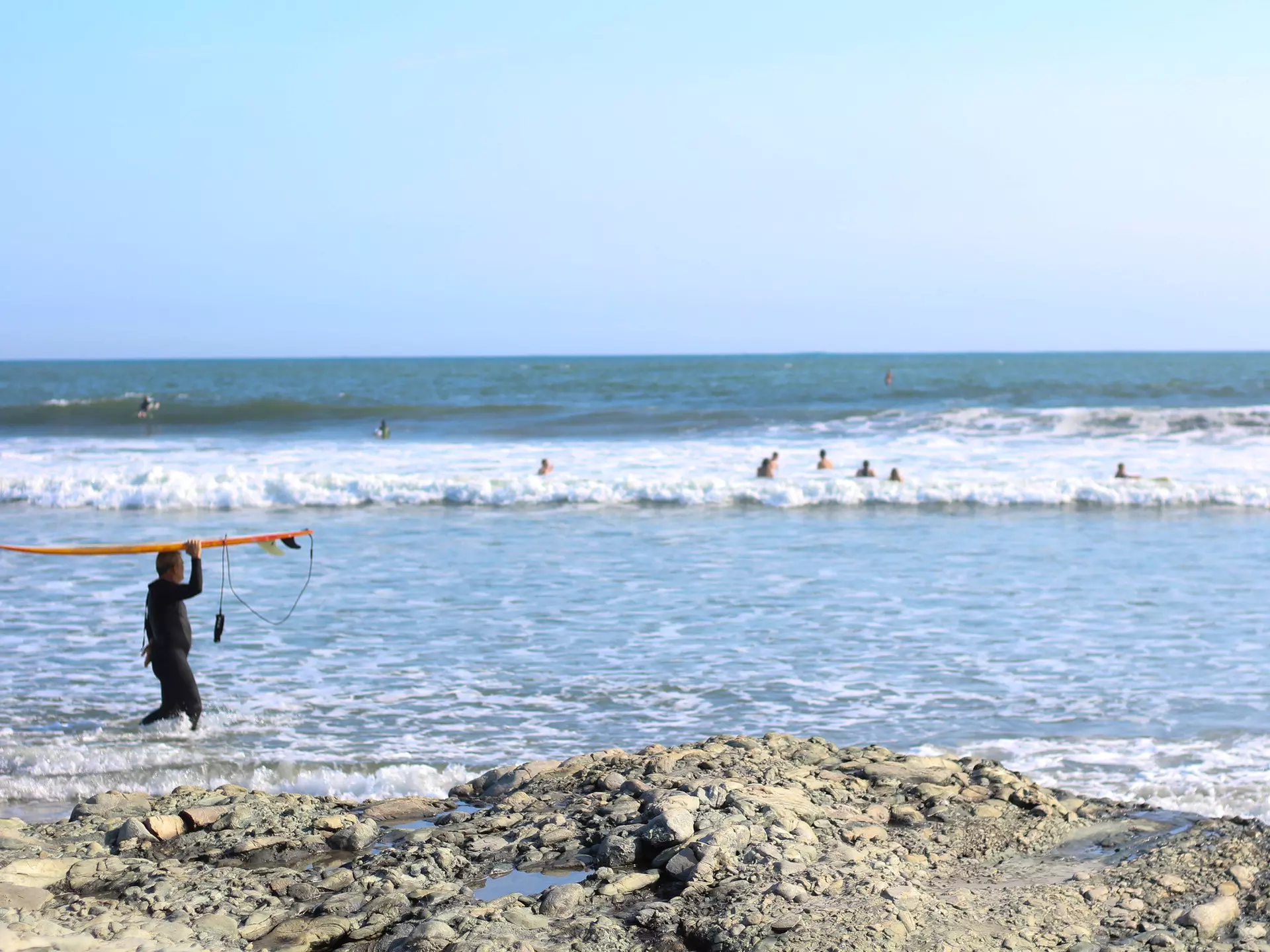 Man in black wetsuit carrying a surfboard above his head walking into the surf with swimmers and the ocean in the background.