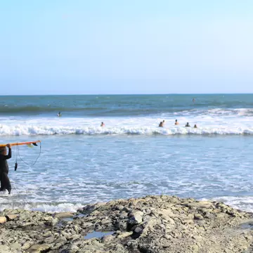 Man in black wetsuit carrying a surfboard above his head walking into the surf with swimmers and the ocean in the background.