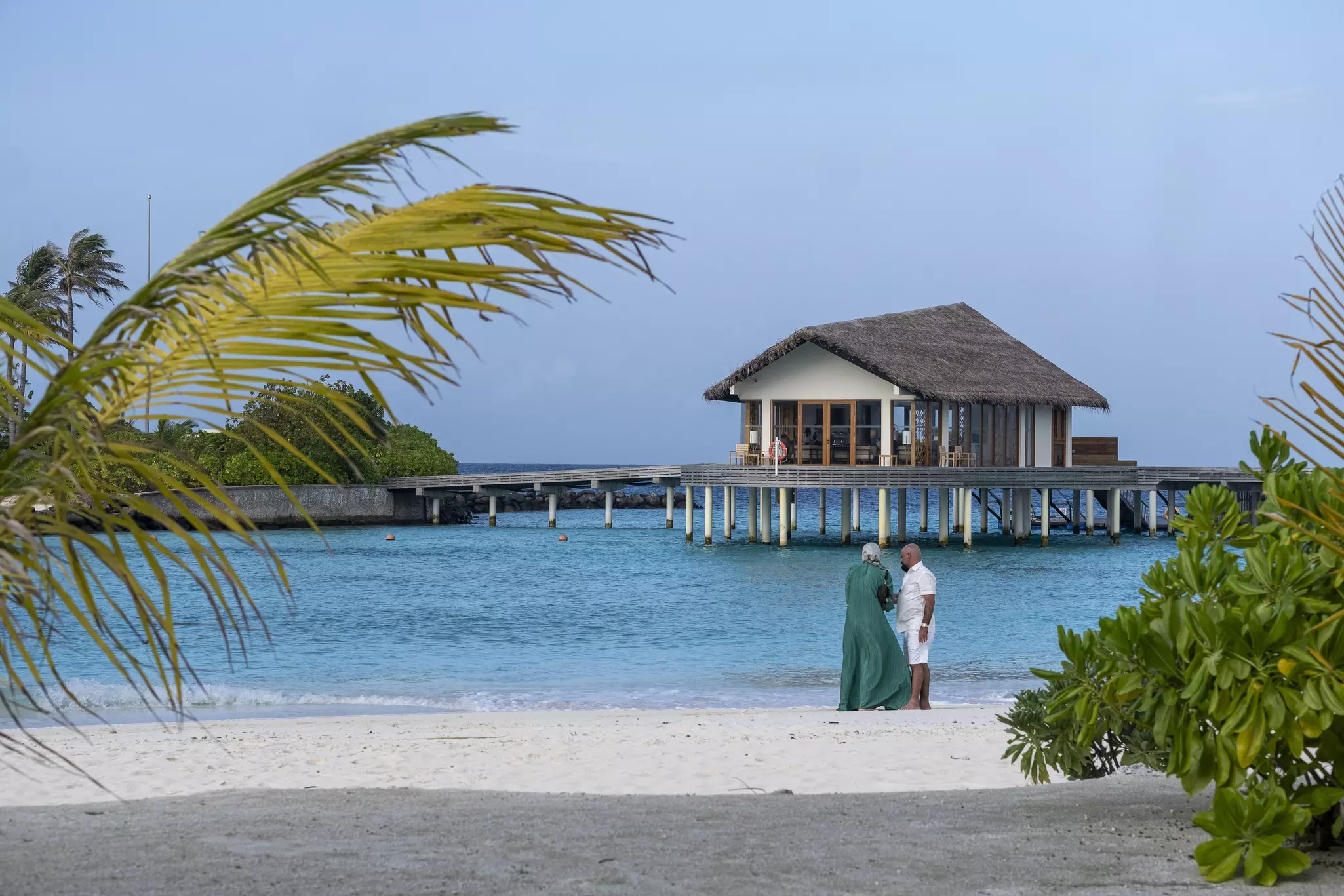 A couple stands on a white sand beach by blue water; a villa is built on stilts over the water. Palm fronds and green bushes are on either side of the sand.