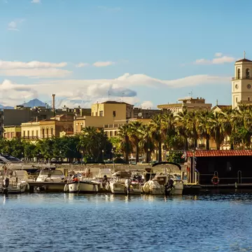 Small motorboats moored along a harbour in Milazzo, Sicily, backed by palm trees, a clock tower and distant mountains.