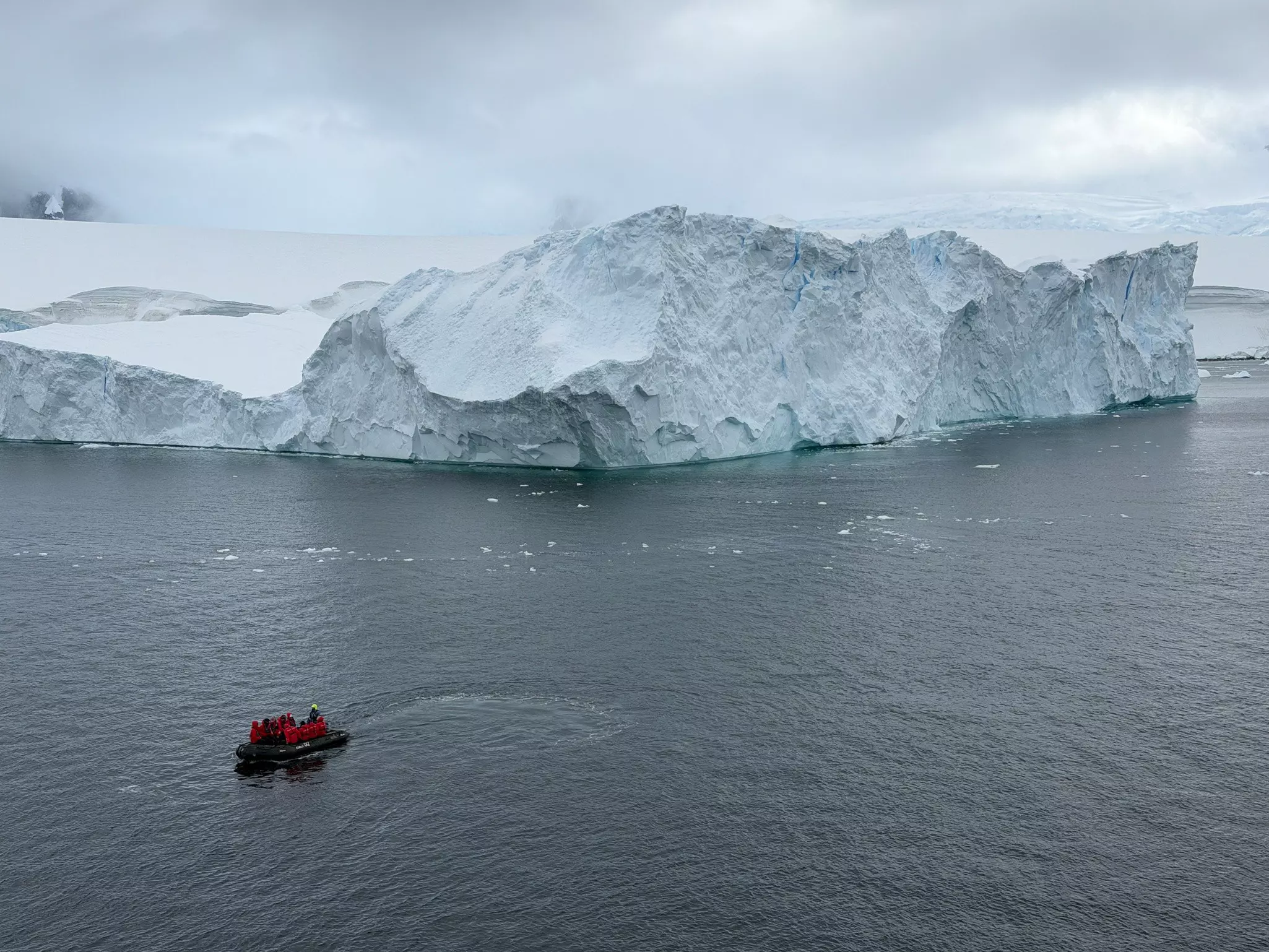 A small boat with people in red jackets approach a massive ice sheet across dark water.