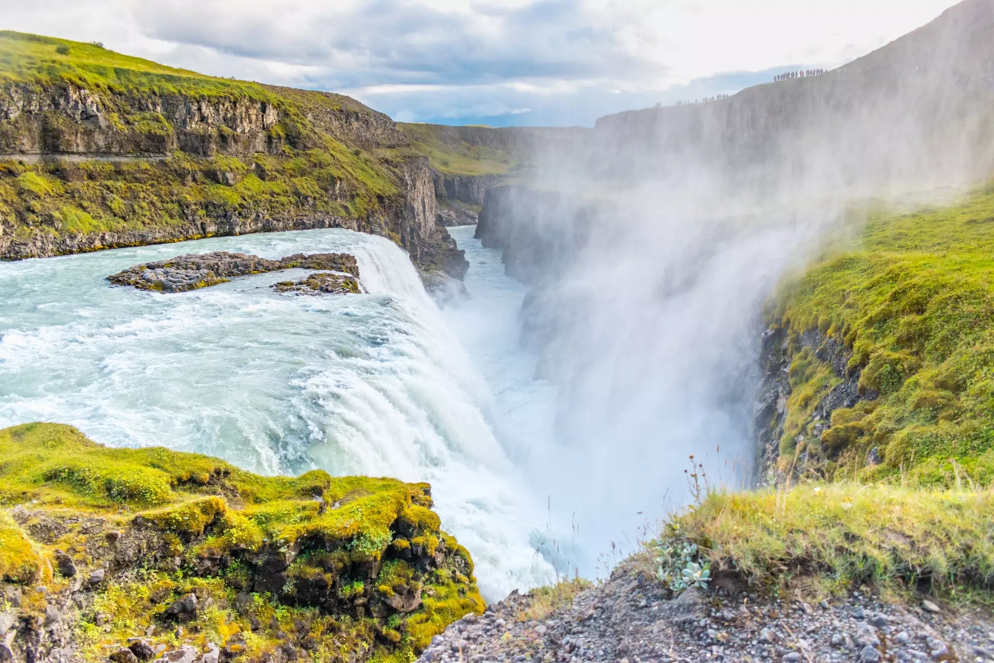 Spray rises from a a huge waterfall that plunges into a ravine, surrounded by green grass and moss.
