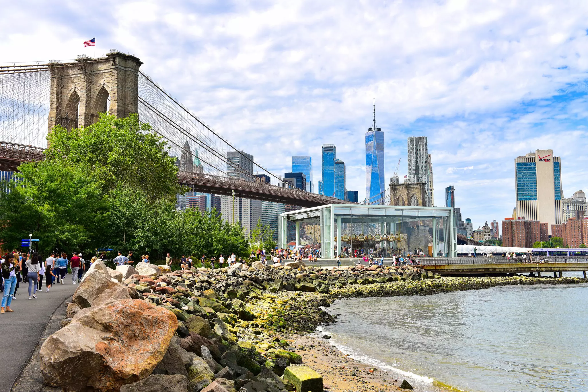 A waterfront park with a large carousel in the shadow of a suspension bridge with brick towers.