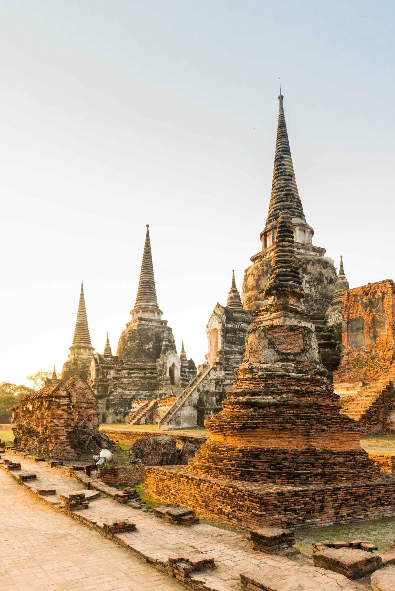 Tall pointed stone stupas and crumbling red-brick ruins beside a brick walkway, lit by warm early-morning sun.