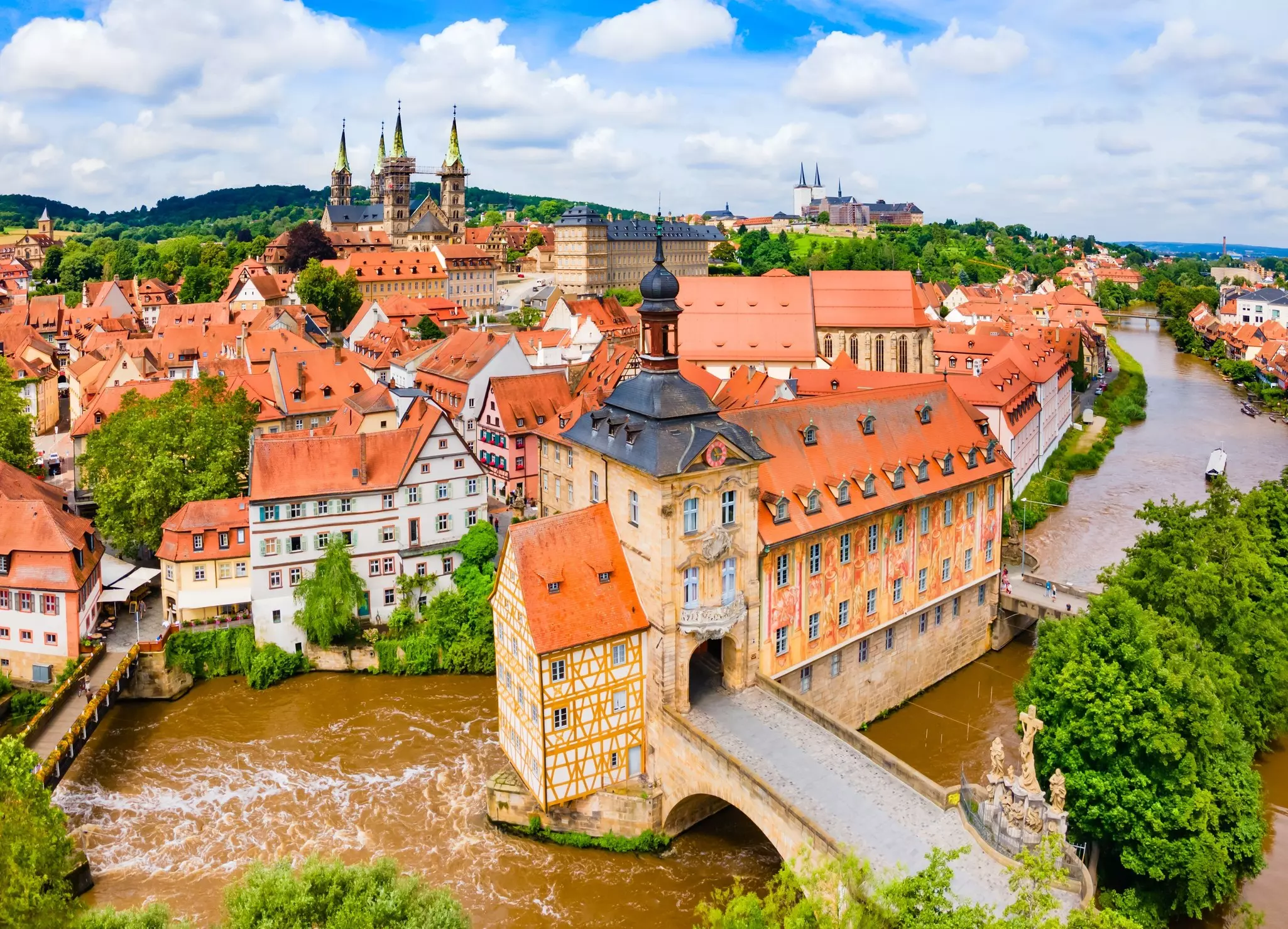 An aerial view of red-roofed buildings in a historic town. A bridge spans a narrow river in the foreground.