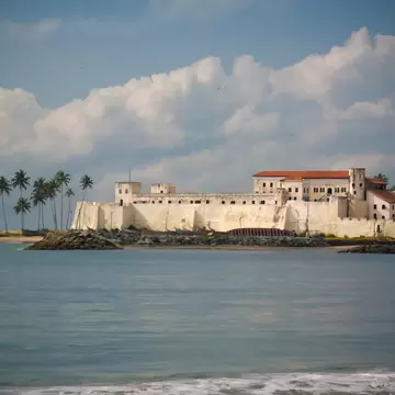 A long whitewashed fortress with red-tiled roofs and a battlemented seaward wall standing on a low headland, palms behind and surf breaking in front.