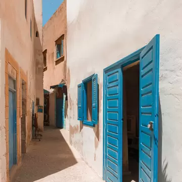 A narrow whitewashed alley of lime-rendered walls, with bright blue shutters and a pair of open blue doors, running back into shade over cobbles.