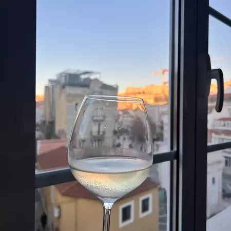 A clear glass of white wine in front of a window looking out at a city at sunset.