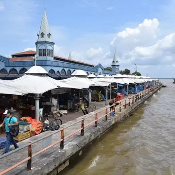 Blue market building with pointed turrets and white-canopied stalls along a quay beside a wide brown river in Belem, Brazil.