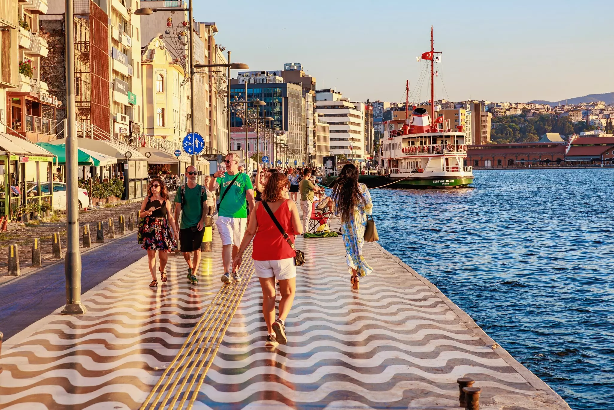 People walk along a waterfront promenade paved with wave-patterned tiles in a major city.