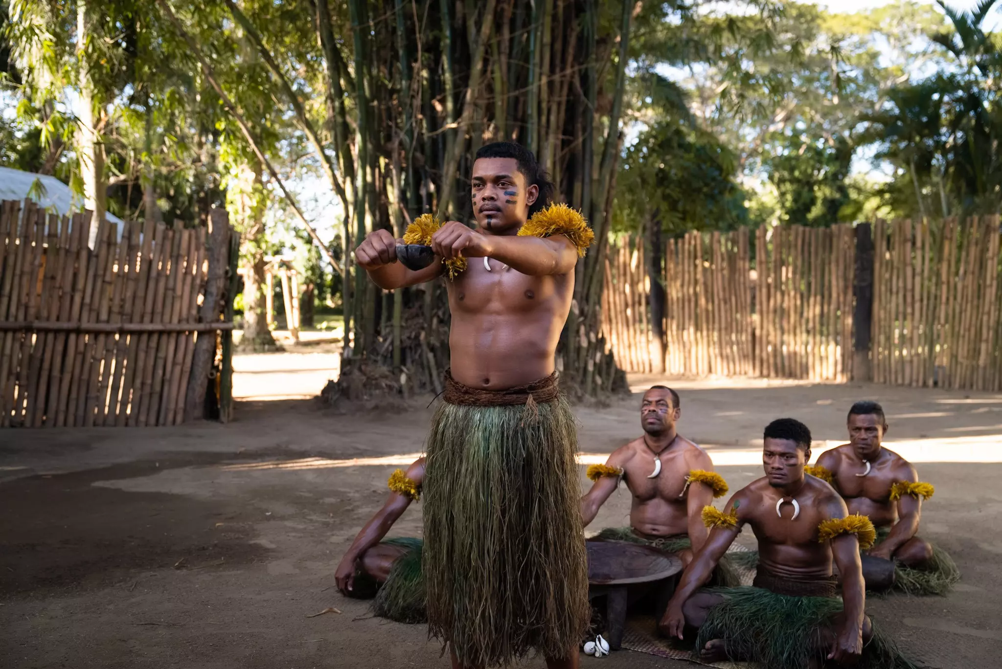 Men in grass skirts seated on the ground with one person standing and holding a cup of kava during a kava ceremony