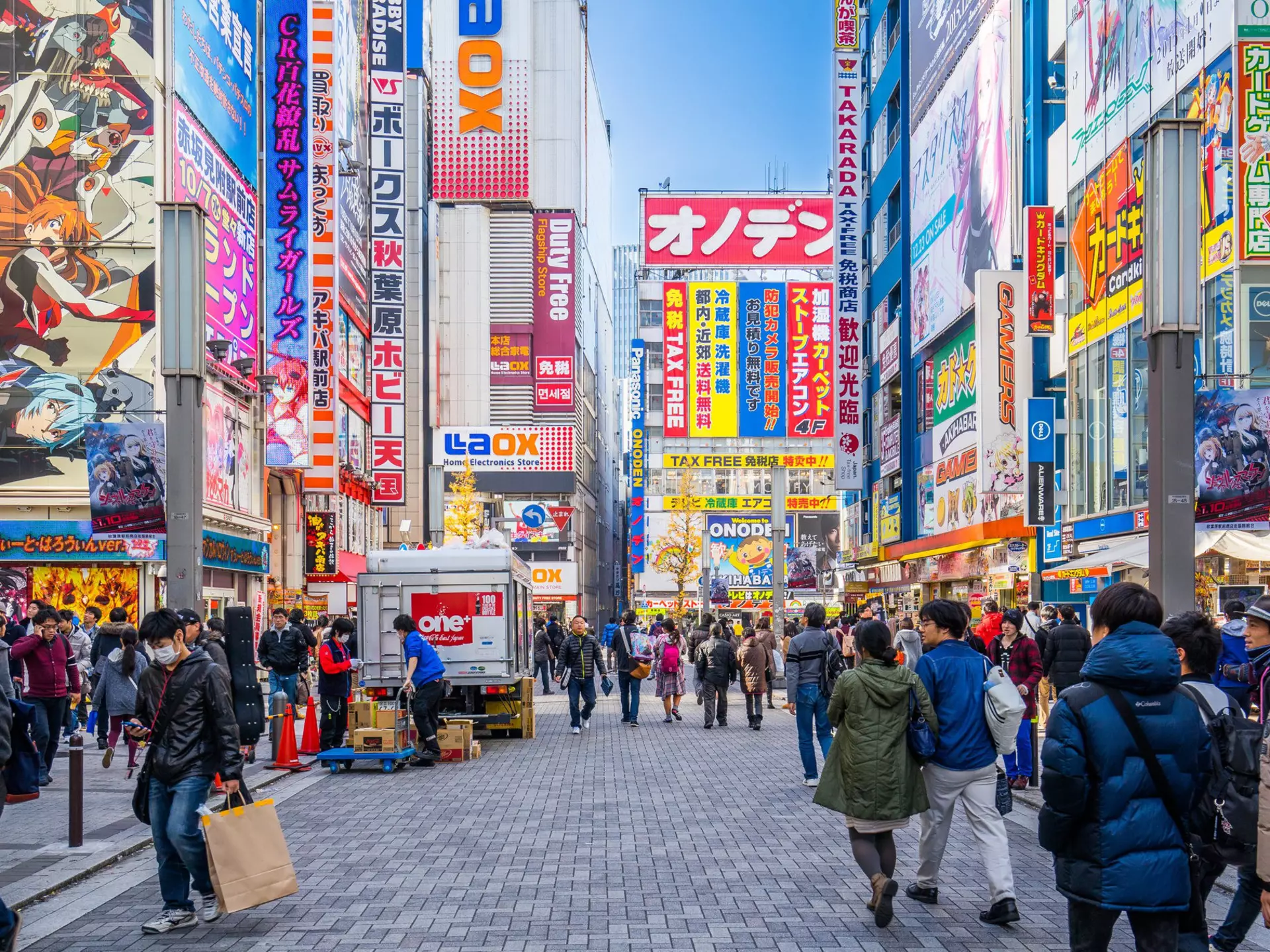 Shoppers walk along a narrow street in Akihabara, tall buildings covered with bright signs on either side