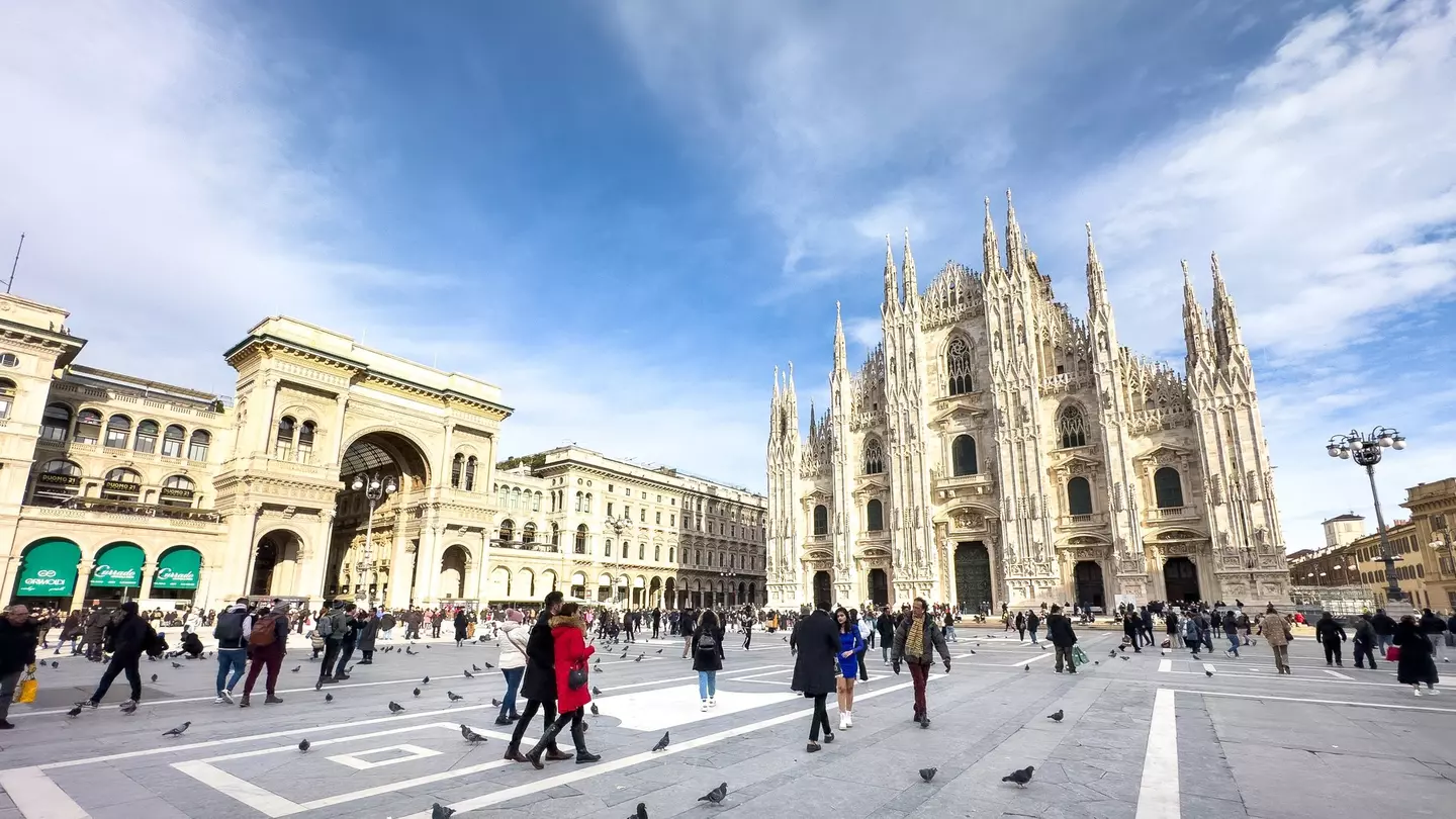 A wide shot of people walking in piazza in a city. An elaborate Gothic church is at one end of the piazza.