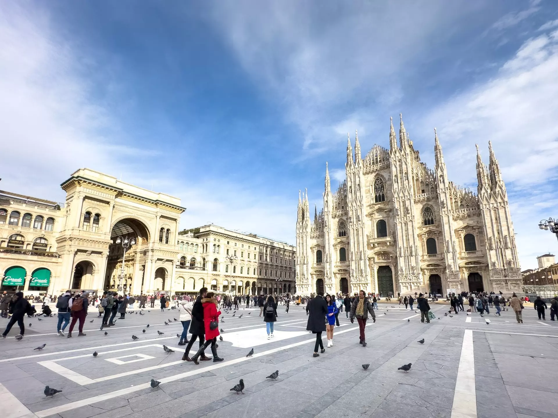 A wide shot of people walking in piazza in a city. An elaborate Gothic church is at one end of the piazza.