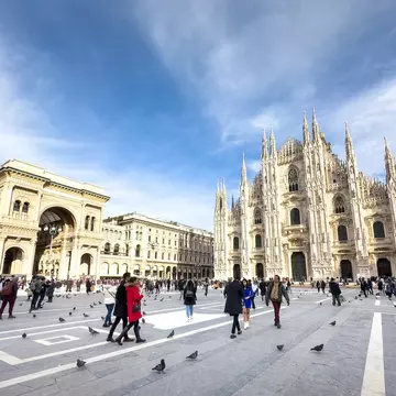 A wide shot of people walking in piazza in a city. An elaborate Gothic church is at one end of the piazza.
