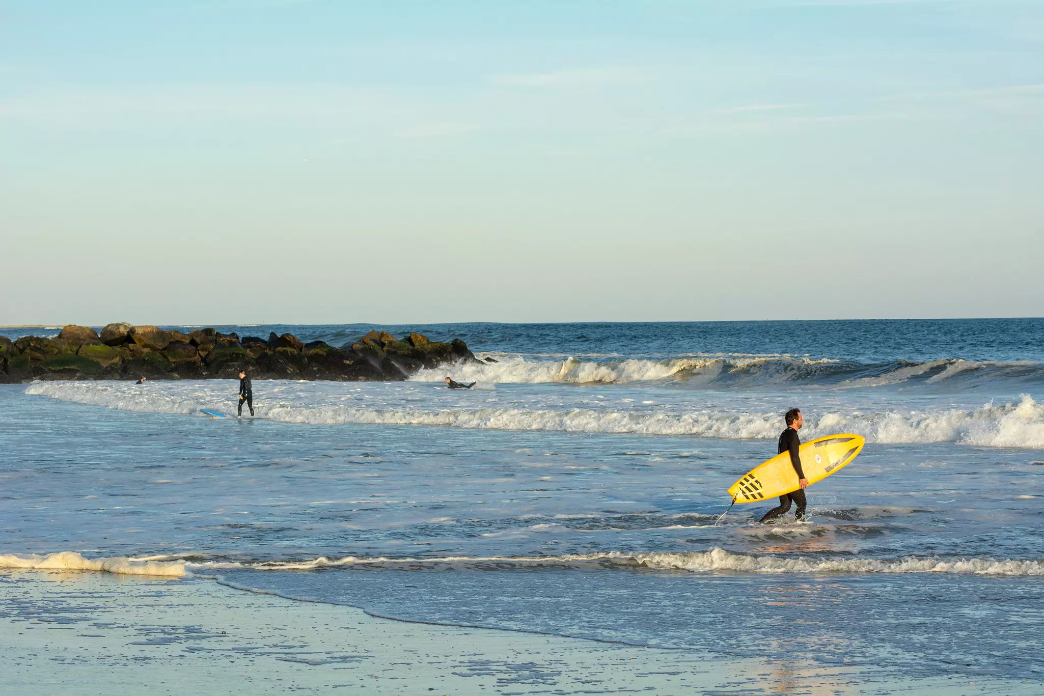 A surfer with a yellow board walks into water with breaking waves.