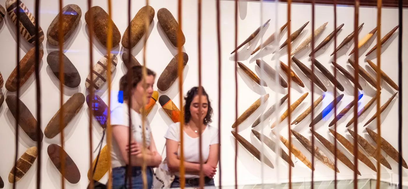 Two museum visitors looking at an exhibit case, with First Nations works displayed on the wall behind them.
