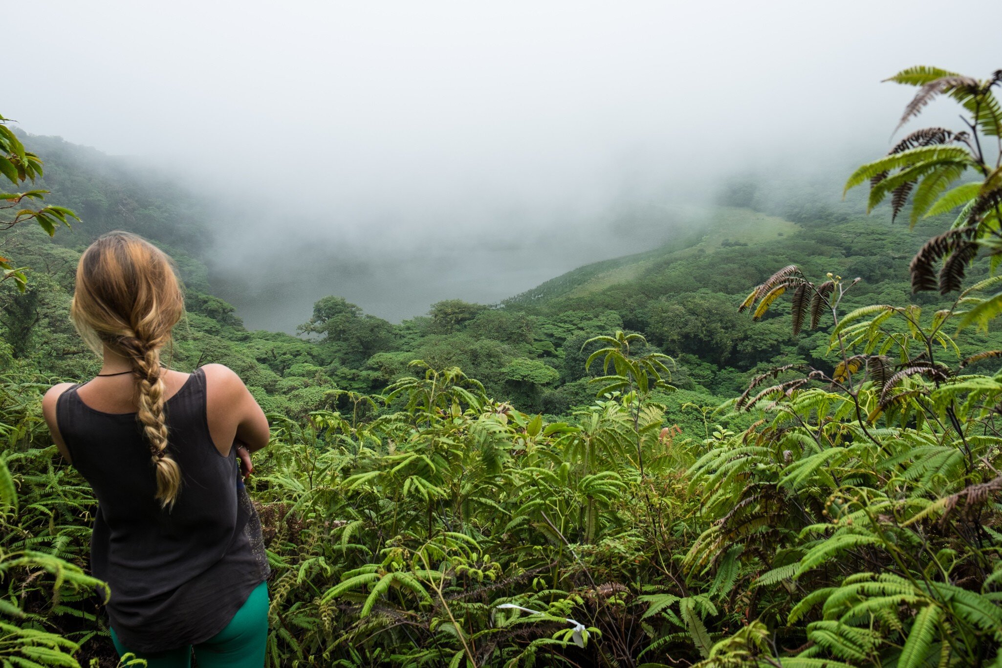 Hiker with a long braid looks over fern-covered slopes toward a cloud-shrouded lake in a forested crater