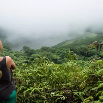 Hiker with a long braid looks over fern-covered slopes toward a cloud-shrouded lake in a forested crater