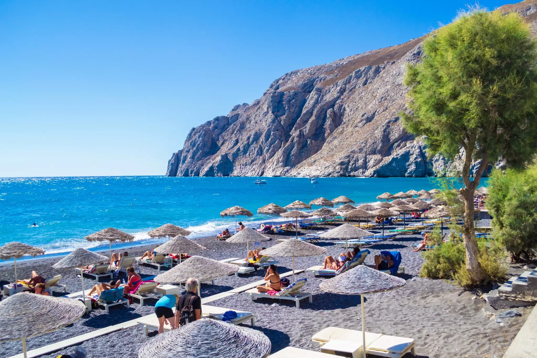 People relax under straw parasols on a black sand beach on a sunny day.
