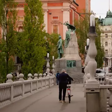 A person walks a bicycle over a bridge toward a statue and a church with a salmon-colored facade.