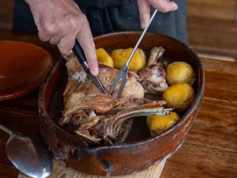 A person's hands hold a two-pronged fork and knife, cutting into a piece of meat surrounded by potatoes in a stew pot.