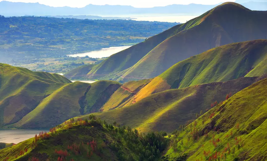 Sunlit green ridges and pine-dotted slopes above the shores of Lake Toba in North Sumatra, with distant hills beyond