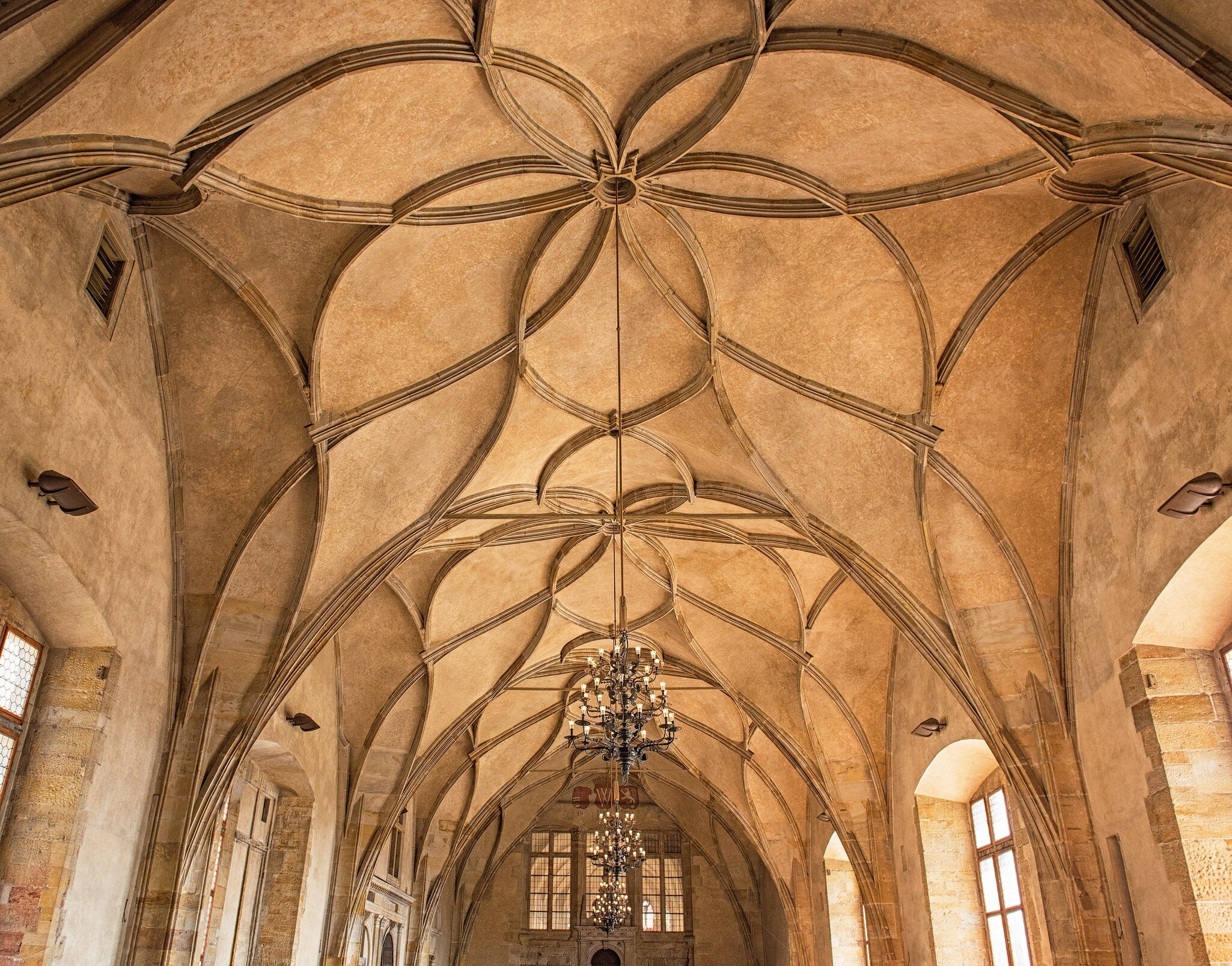 A room with a high, earth-toned ceiling with patterns. Large chandeliers hang in the middle of the room, which has large windows.