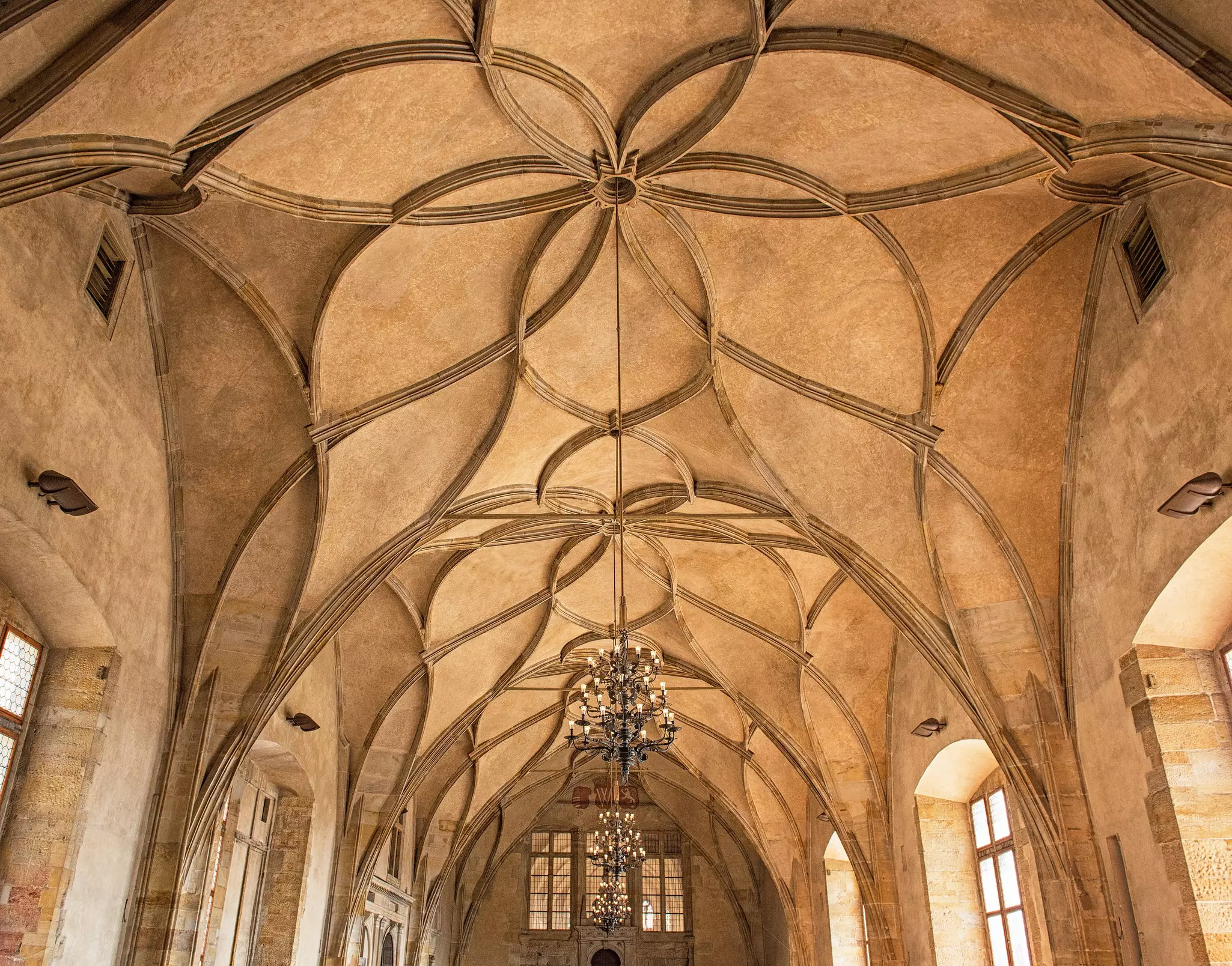 A room with a high, earth-toned ceiling with patterns. Large chandeliers hang in the middle of the room, which has large windows.