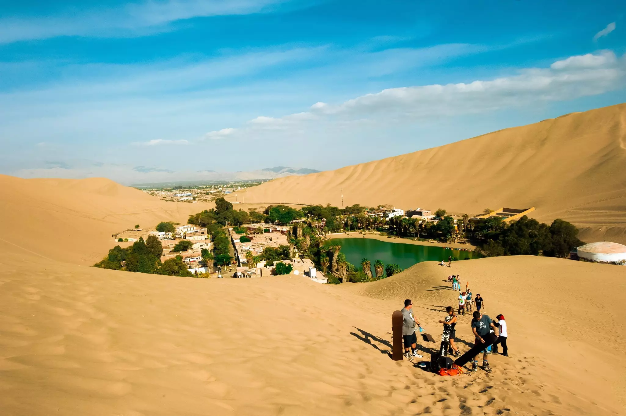 A small town surrounded by steeps sand dunes. A group of people prepare to sandboard down the dunes.