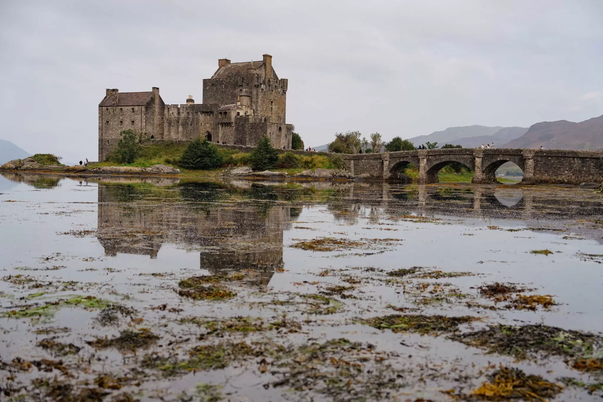 A stone castle with an arched bridge sitting beside a lake on a cloudy day