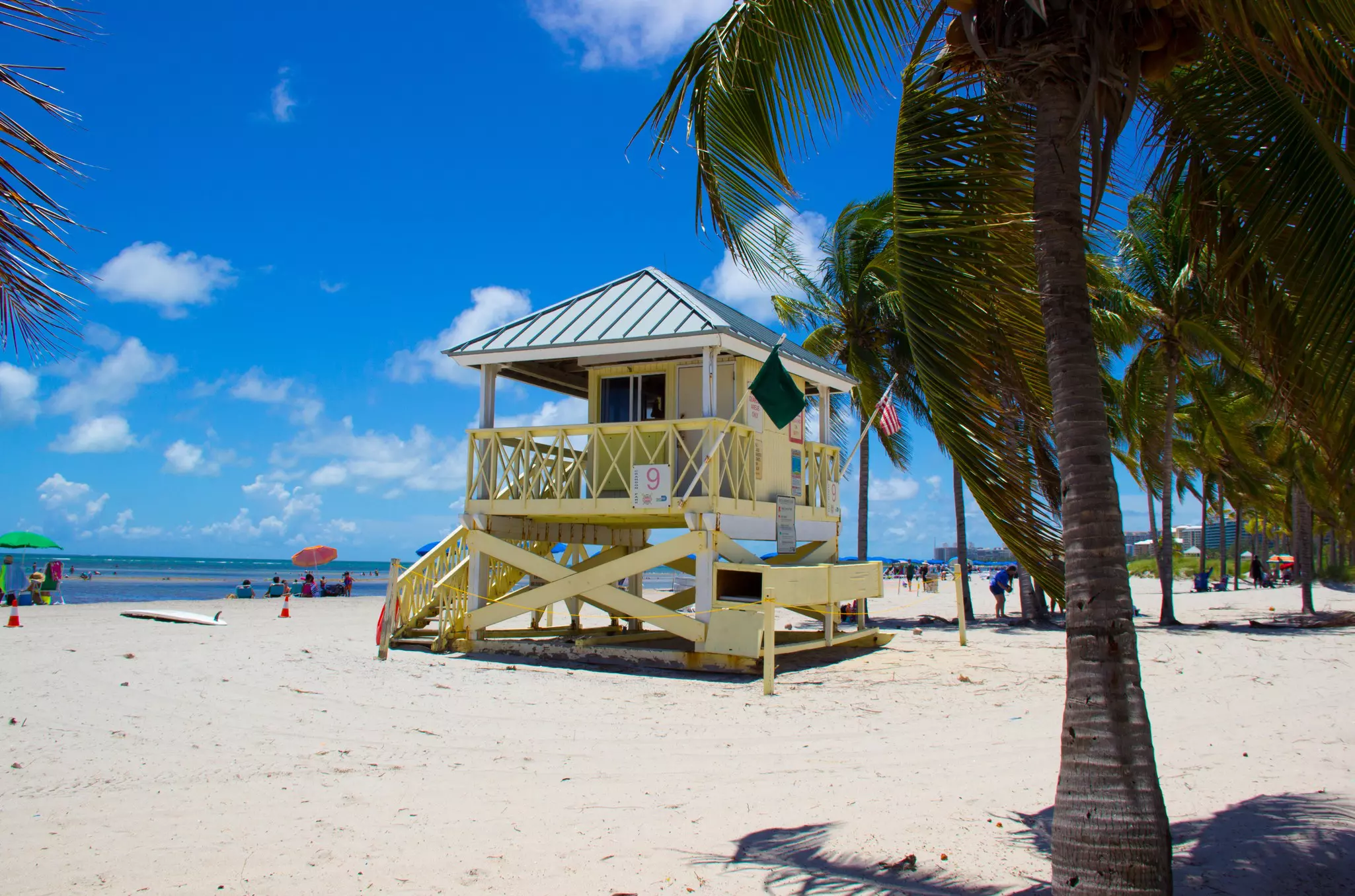 Lifeguard house on a white, sandy beach with palm trees in the foreground and beachgoers and the ocean in the distance.