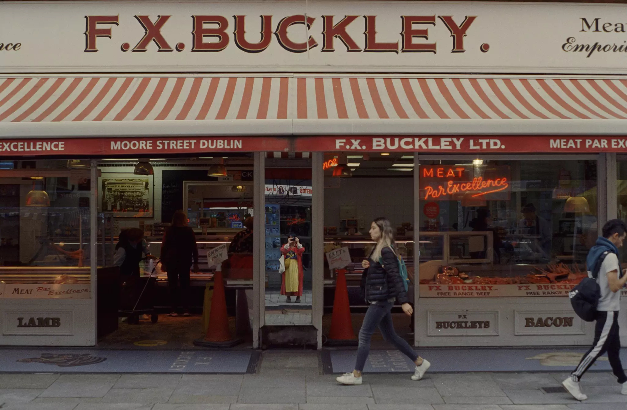 Two people walk past a meat store called F.X. Buckley while the photographer is reflected in a mirror