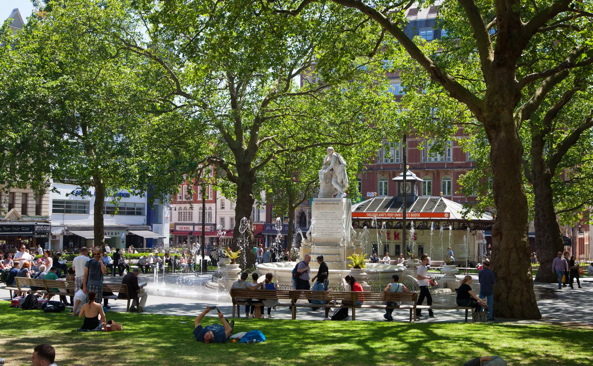 People sit on benches and green grass in a shady park with a statue at the center.
