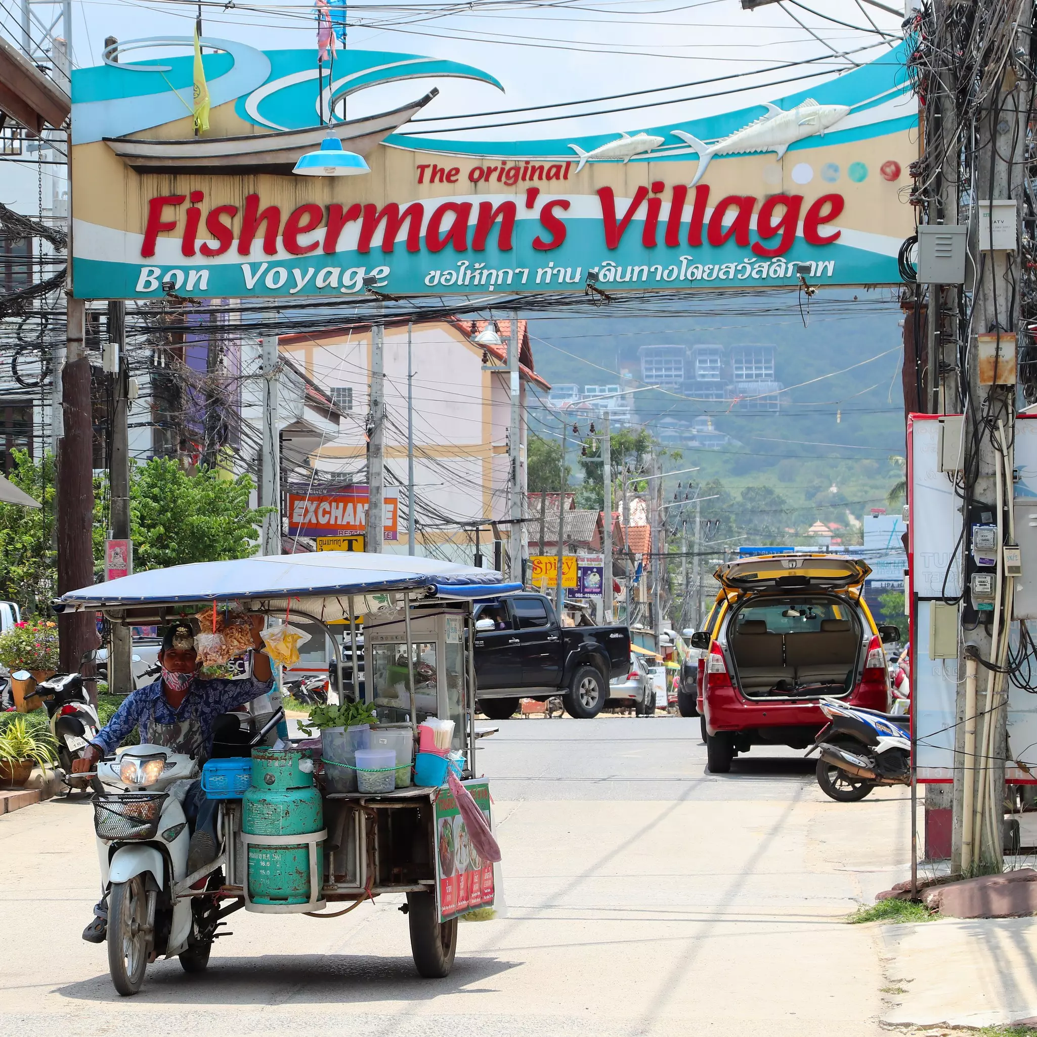 A food vendor with a cart attached to a motorcycle drives a street with a huge sign above it that says "The original Fisherman's Village, bon voyage"