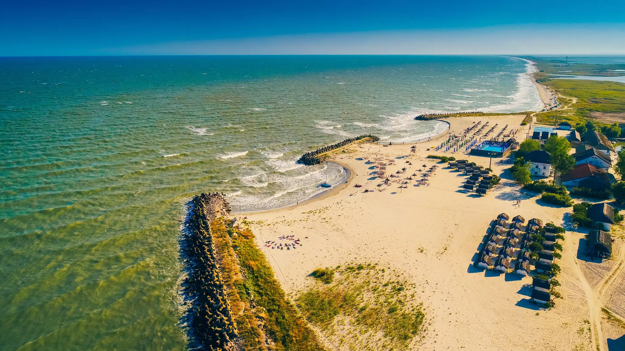 Aerial view of sandy beach bordered by greenery with umbrellas and small structures and a green-colored sea to the left on a sunny day.