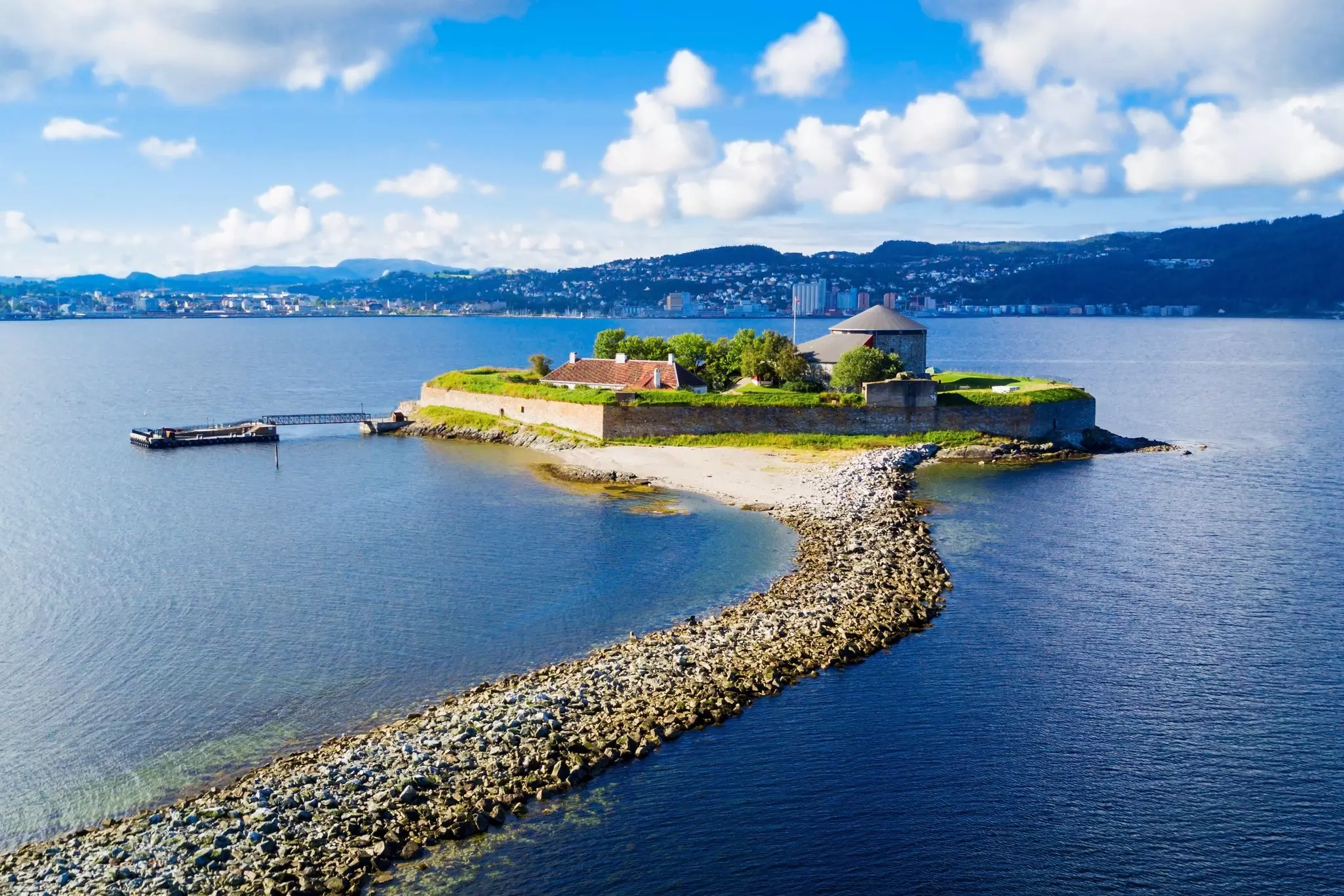 An aerial view of an island with a historic structure in the harbor of a city. A breakwater is seen in the foreground of the image.